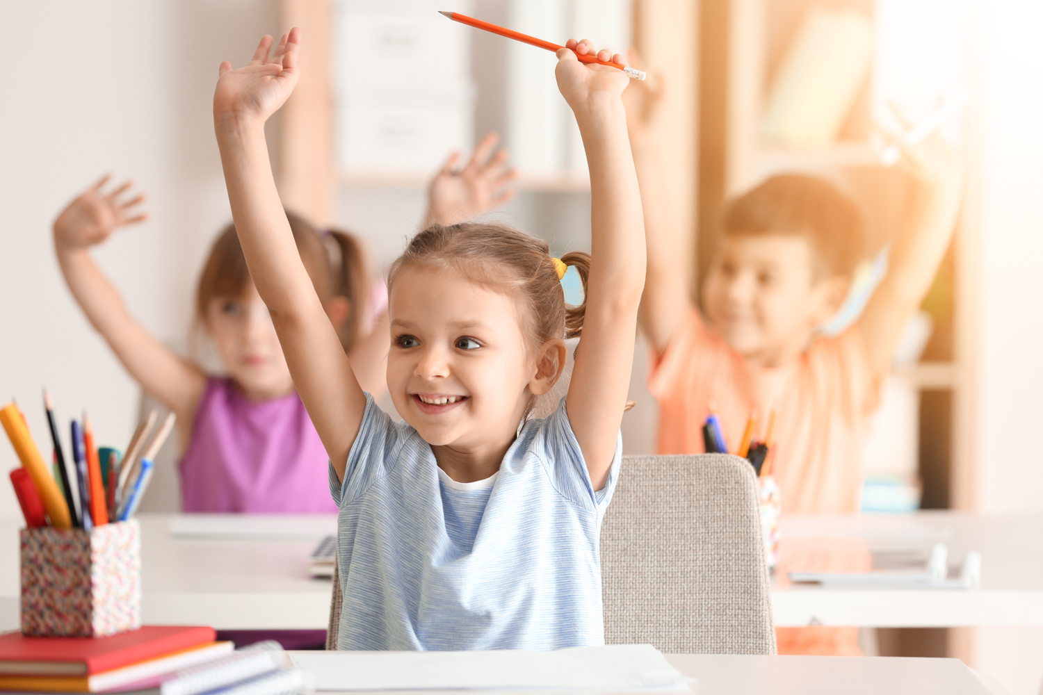 På bilden syns tre barn som sträcker upp händerna i luften samtidigt som de ler sittandes vid sina bänkar i ett klassrum.